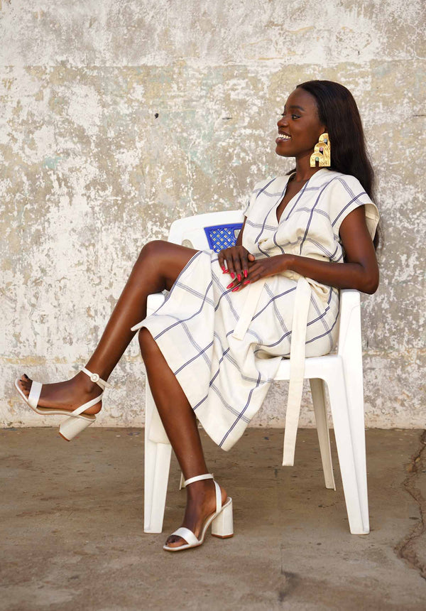 Woman sitting on a white chair wearing a white dress with blue patterns and white sandals.